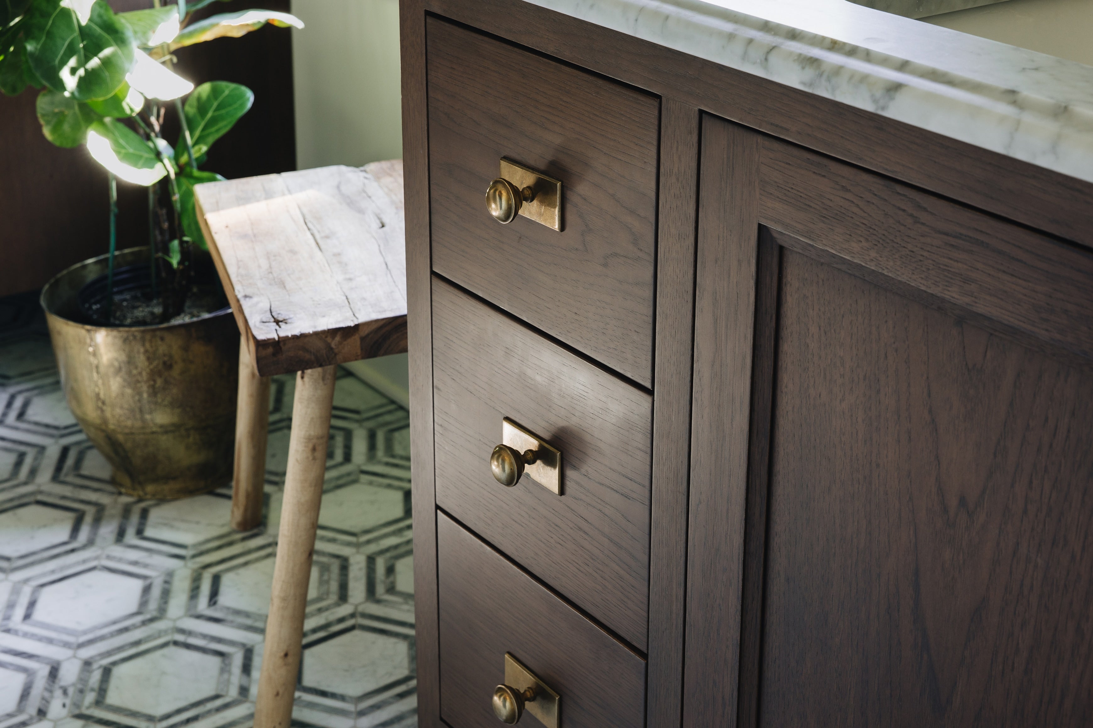 Dark brown wooden bathroom vanity cabinet with three drawers and brass round knobs, shown next to a marble countertop and a potted plant on a patterned tile floor.