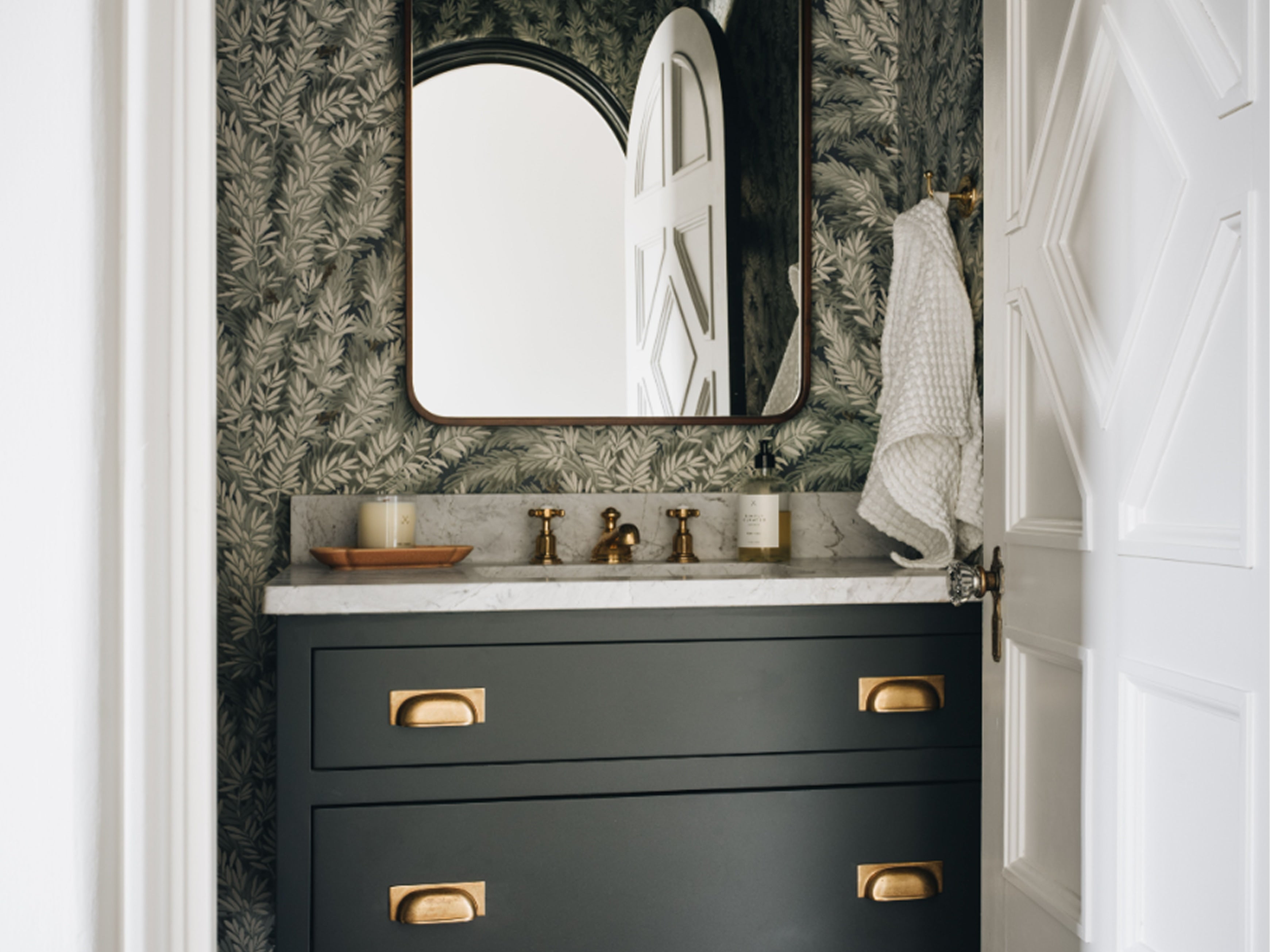 A modern bathroom vanity featuring a matte black cabinet with brass handles, a white marble countertop, a matching wall mirror, and dark patterned wallpaper. A white textured towel hangs on the right side.