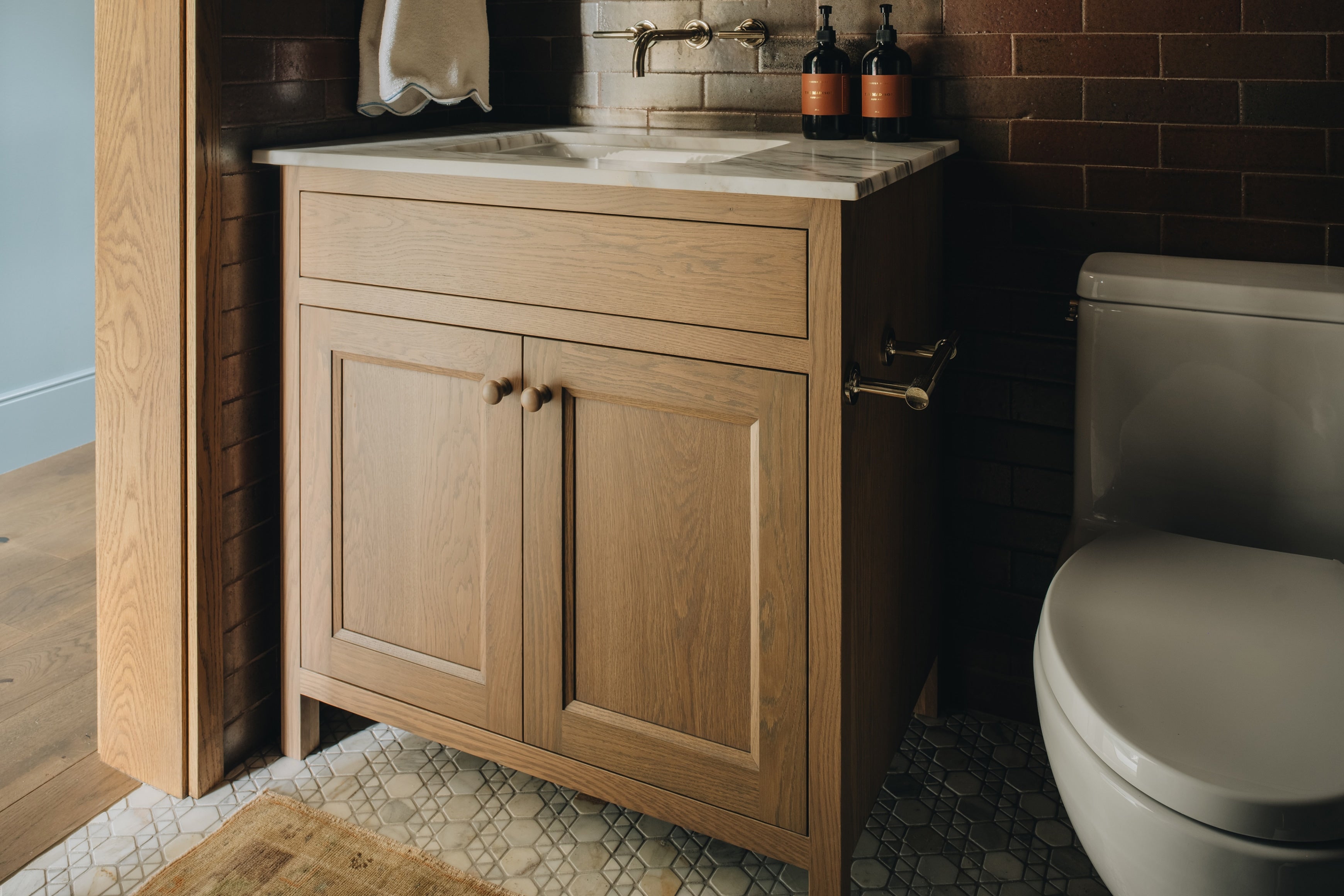 A light oak wood bathroom vanity cabinet with a white stone countertop and integrated sink, shown in a styled bathroom setting next to a toilet and against a dark brick wall.