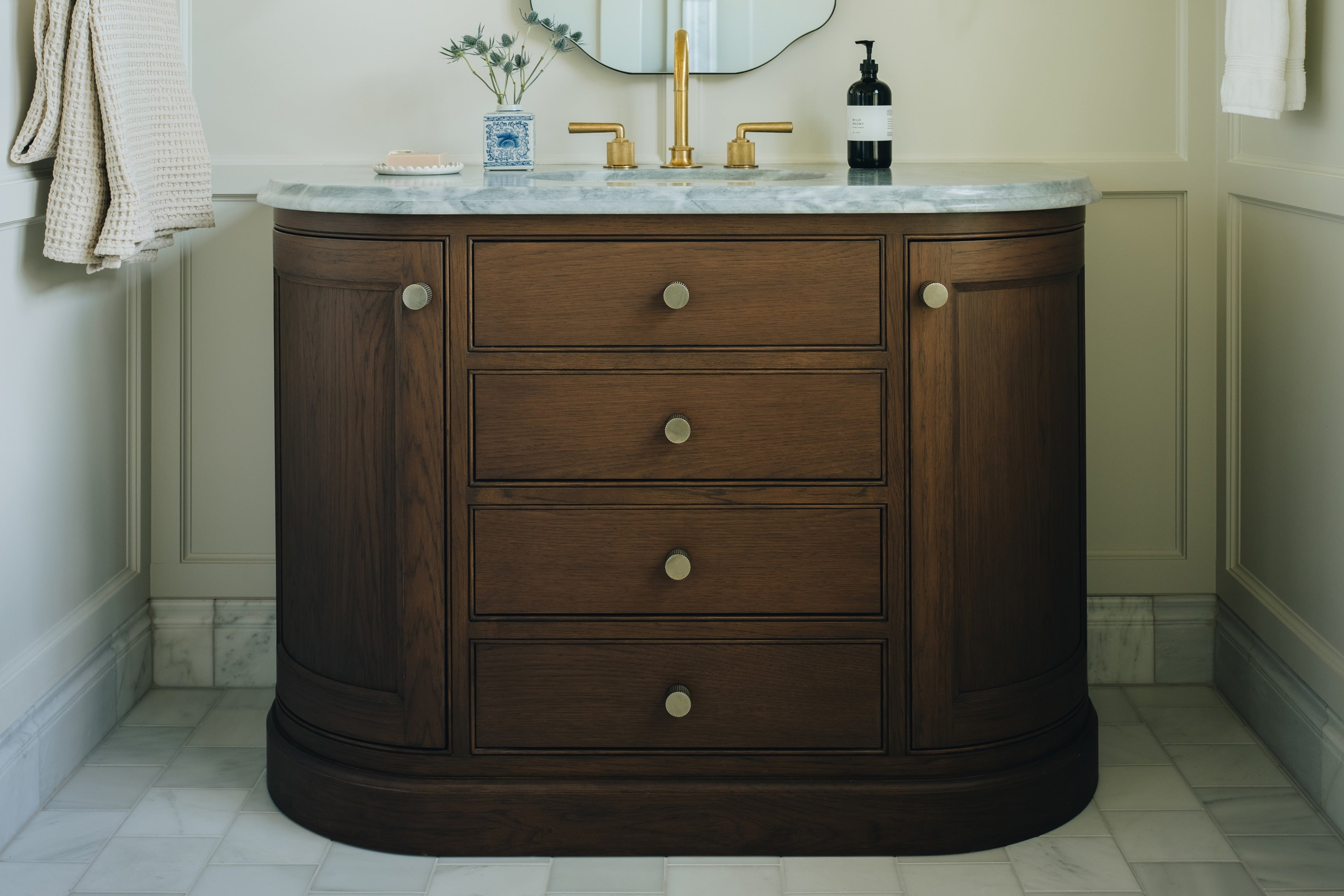 A curved dark brown wooden bathroom vanity with four drawers and brass knobs, topped with a white marble countertop and gold faucet, set in a classic bathroom interior.
