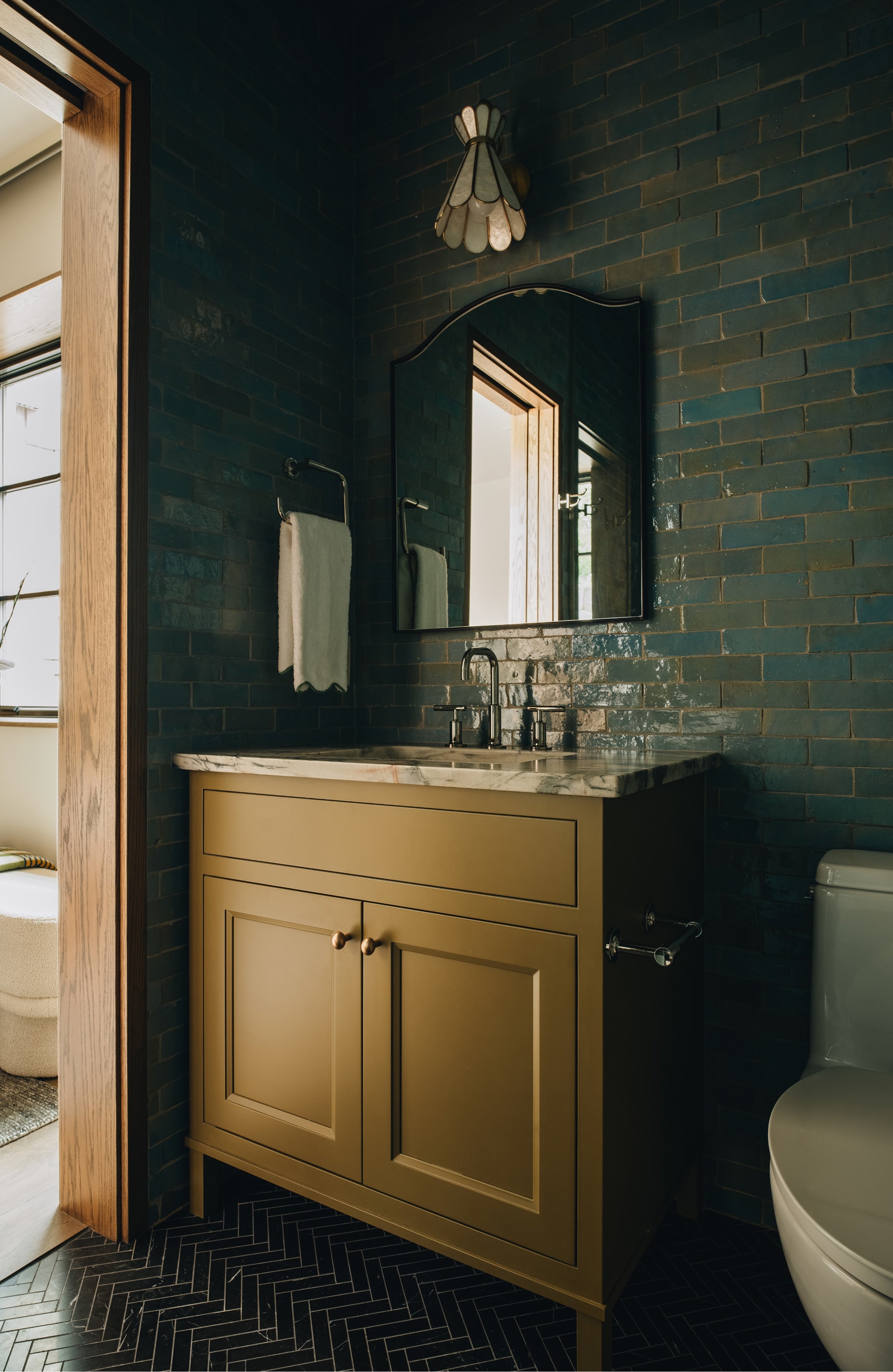 A vintage-style bathroom vanity with a mustard yellow cabinet and a marble countertop, set against a dark teal brick wall with a matching mirror.