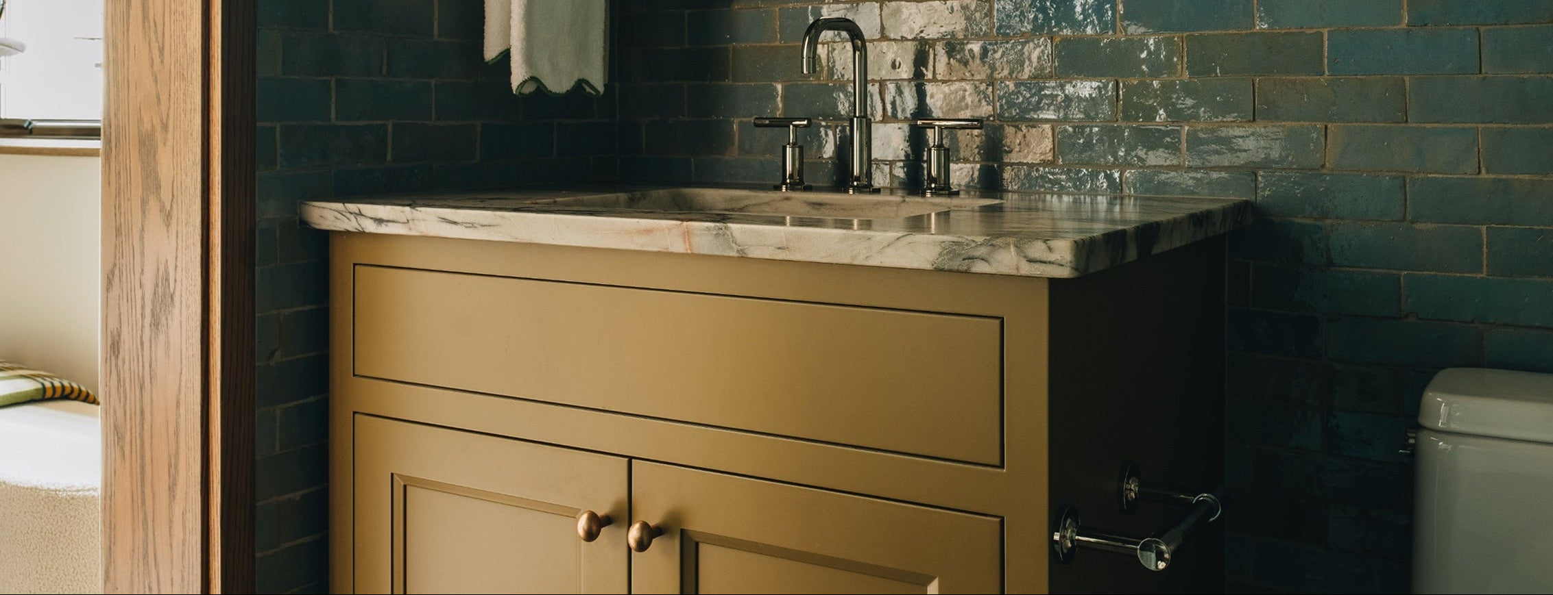 A vintage-style bathroom vanity with a mustard yellow cabinet and a marble countertop, set against a dark teal brick wall with a matching mirror.