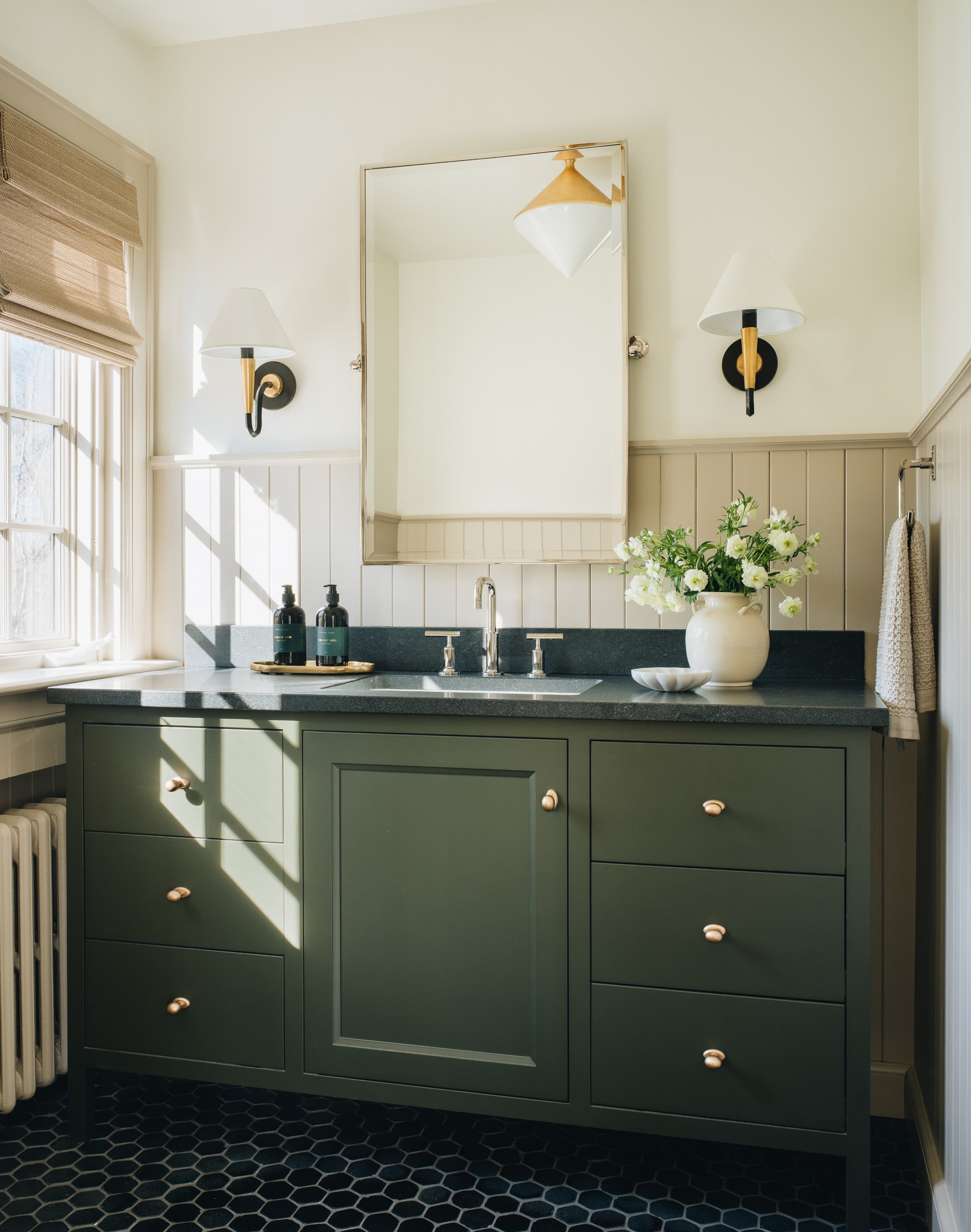 A dark green bathroom vanity cabinet with brass knobs, a black countertop, and an undermount sink, shown in a styled bathroom setting with a mirror, wall sconces, a window, and a floral arrangement.